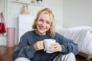 Portrait of young woman sitting on bedroom floor, drinking tea, holding white mug and smiling at camera photo