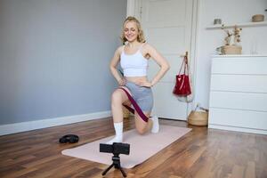 Portrait of young athletic woman recording home workout , shooting content for sport fitness vlog, using resistance band and digital camera photo