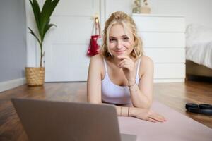 retrato de linda joven aptitud mujer, haciendo rutina de ejercicio a hogar, acostado en caucho yoga estera, acecho ejercicios en línea en ordenador portátil foto