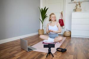 retrato de joven aptitud bloguero, mujer demostración ejercicios a su seguidores, grabación en digital cámara, haciendo rutina de ejercicio formación sesión, hogar yoga, sentado en frente de ordenador portátil en caucho estera foto