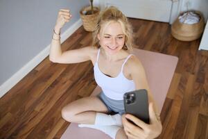 retrato de joven y ajuste aptitud mujer, formación instructor sentado en un habitación a hogar, utilizando yoga estera, flexionando bíceps, ejercicio, grabación sí misma durante ejercicios foto