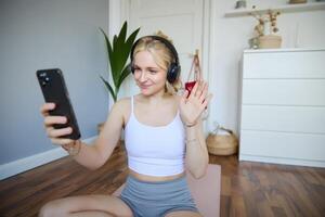 Portrait of young sporty woman in headphones, records on her smartphone, live streaming and saying hello to followers while doing workout training at home photo