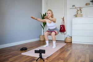 Portrait of woman, fitness instructor at home, recording about workout, showing how to do leg exercises, squats with elastic resistance band, working out indoors on yoga mat photo