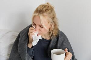 Concept of flu and quarantine. Close up portrait of young woman feeling sick, caught a cold, sneezing and blowing nose in napkin, holding warm tea in hand photo