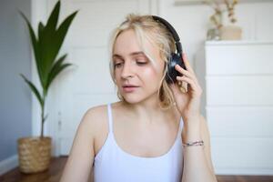 Portrait of good-looking young blond woman in wireless headphones, listens to podcast or favourite song, enjoys music in new earphones photo