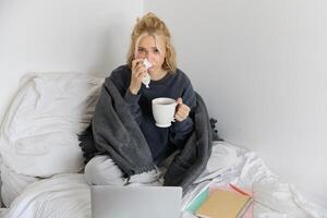 Portrait of young woman sitting at home sick, feeling unwell, drinking tea, spending time in bed, has runny nose, working on laptop photo