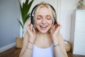 cerca arriba retrato de mujer sonriente mientras escuchando a música en inalámbrico auriculares, canto con ojos cerrado foto