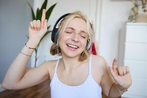 Portrait of happy and carefree blond woman, listens to music in wireless headphones, dancing and singing along favourite song, spending time alone at home photo