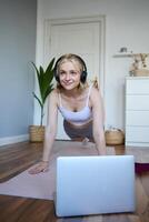 Vertical shot of young blond woman at home, wearing wireless headphones, looking at laptop, standing in plank, watching fitness tutorial online photo