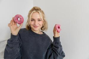 Close up portrait of happy, cute blond woman, holding doughnut, eating sweet, delicious comfort food, showing dessert at camera photo