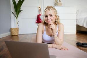 Portrait of young fitness woman, doing workout at home, looking at her gym instructor on laptop screen, doing online training session photo