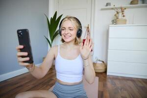 Portrait of young sporty woman in headphones, records on her smartphone, live streaming and saying hello to followers while doing workout training at home photo