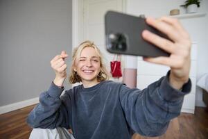 Portrait of young stylish girl sits on bedroom floor, takes selfies on her smartphone, posing for photo on social media app, smiling and looking happy at camera