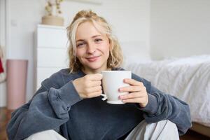 Portrait of young woman sitting on bedroom floor, drinking tea, holding white mug and smiling at camera photo