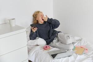 Portrait of young woman spending lazy weekend at home, lying in bed with smartphone and laptop, eats doughnut and yawns, feels sleepy or bored photo