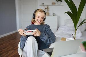 Portrait of smiling young woman, watching tv show in headphones, eating breakfast and looking at laptop screen photo