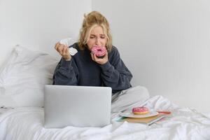 retrato de triste, llorando joven mujer, quedarse a hogar, sentado en cama con rosquilla y comodidad alimento, mirando a alguna cosa molesto en ordenador portátil pantalla foto