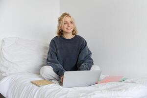 Portrait of young smiling woman studying in her bed, working from home in bedroom, sitting with laptop and notebooks on lotus pose, looking happy and relaxed photo