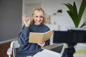 Portrait of smiling young woman, holding notebook, showing okay, ok sign, looking at digital camera, recording , creating content photo