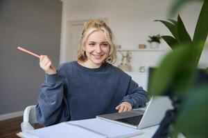 Portrait of young charismatic woman, smiling, holding pen, looking at laptop, chats, doing online course, answering a question, studying with tutor photo