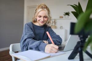 retrato de joven mujer, estilo de vida bloguero, grabación de sí misma, haciendo notas, escritura en diario, sentado en frente de ordenador portátil en un habitación y estudiando foto