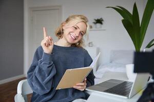 Portrait of young smiling, charismatic woman, raising one finger, teaching online, reading from notebook, looking at laptop, recording course, content maker working on new project from home photo