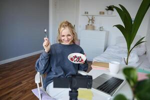 Portrait of cute young girl vlogger, showing her breakfast on camera, recording vlog in her bedroom, holding dessert, eating photo