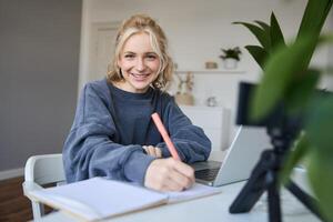 linda sonriente niña se sienta en un habitación, escribe abajo notas, haciendo tarea, registros de sí misma en digital cámara, crea contenido para vlog, estilo de vida blogger haciendo diario rutina episodio foto