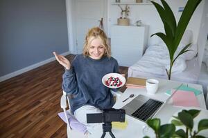 Portrait of young woman talking to audience, recording vlog on digital camera, showing her breakfast, talking about healthy food and lifestyle photo