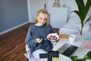 Portrait of smiling young female vlogger, recording and creating content in her room, eating in front of camera, talking, making lifestyle blog photo