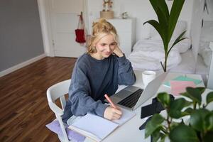 Portrait of smiling, beautiful blond woman, writing down notes, doing homework, studying from home, doing distance learning, online course, working remotely in her room photo