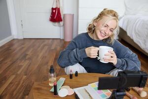 Young woman smiling, recording lifestyle vlog on her digital camera, holding cup of tea while talking, has makeup on coffee table, doing makeup tutorial for social media from her room photo