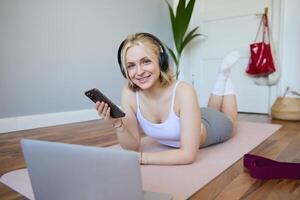 Portrait of young sporty woman, connects to online workout training session, doing exercises, lying on rubber mat, listens to fitness instructor in headphones, holding mobile phone photo