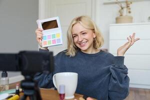 Lifestyle blogger, records in her room, has a camera on coffee table, shows eyeshadow palette to her followers, does makeup tutorial, vlogger working indoors, creates content for social media photo