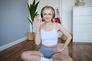 retrato de joven sonriente hermosa mujer, vistiendo inalámbrico auriculares, trabajando fuera a hogar, sentado en caucho yoga estera, demostración bueno firmar, satisfecho con ejercicios foto