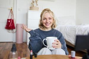 Cheerful woman, beauty blogger, records lifestyle vlog on digital camera, talks casually, tells a story for social media followers, holds cup, drinks tea and sits on floor in her room photo