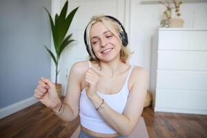 retrato de feliz, hermosa atlético mujer, trabajando fuera a hogar, escuchando música a aumentar energía mientras activo formación sesión, utilizando inalámbrico auriculares y caucho yoga estera foto