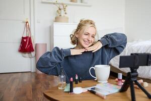 Lifestyle blogger records of herself while doing hairstyle, showing makeup lifehacks for social media followers, vlogging, using camera for her blog, sitting in a room, drinking tea photo