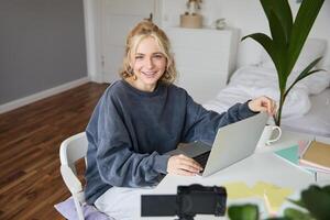 Portrait of stylish, cute young blond woman, sitting with laptop in a room, recording on digital camera, vlogging, making lifestyle content for social media photo