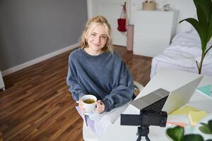 Portrait of young smiling woman, social media influencer, girl with digital camera and laptop, sits with cup of tea in bedroom, records vlog, creates content photo