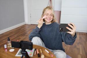 Image of young woman, vlogger taking selfie in her room, talking to her followers during online live stream, using smartphone app to chat with audience, smiling and looking happy photo