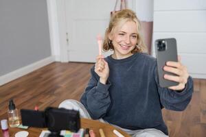 Image of stylish young woman, social media influencer, taking pictures on mobile phone, doing makeup tutorial for followers online, recording vlog in her bedroom, showing brush photo
