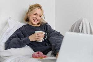 retrato de sincero, contento joven mujer acostado en cama, mirando a ordenador portátil pantalla, participación taza de té y comiendo rosquilla, quedarse a hogar en fin de semana, gasto bastante hora solo foto