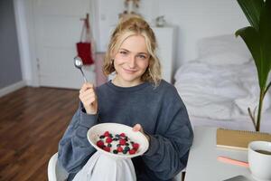 Portrait of cute smiling blond woman eats breakfast in her bedroom, looking at camera, holding bowl with dessert and spoon photo