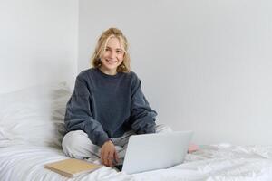 Portrait of young smiling woman studying in her bed, working from home in bedroom, sitting with laptop and notebooks on lotus pose, looking happy and relaxed photo