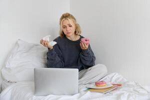 Portrait of sad, crying young woman, staying at home, sitting in bed with doughnut and comfort food, looking at something upsetting on laptop screen photo