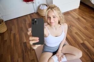 Young sporty fitness trainer, yoga instructor does live stream, takes selfies on smartphone while sitting on rubber mat at home, recording her workout training session photo