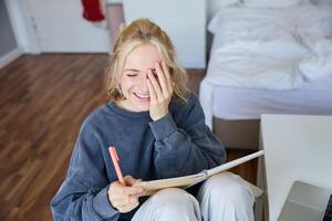 retrato de carismático rubio chica, sonriente mujer en dormitorio, participación cuaderno y bolígrafo, escritura en diario o diario, crea a hacer lista en planificador foto