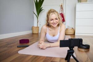 Portrait of young woman, sports vlogger, fitness instructor recording of herself showing workout exercises, using digital camera, lying on yoga rubber mat photo