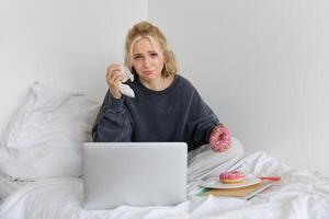 Portrait of woman watching sad movie on laptop, crying and wiping tears off, eating doughnut and drinking tea photo